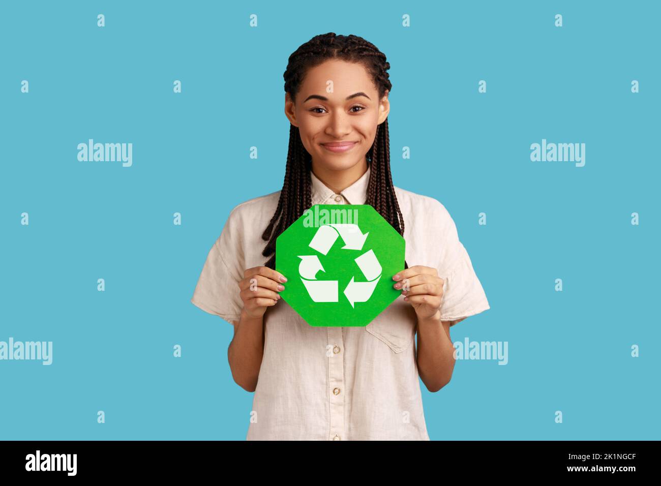 Responsible self confident woman with black dreadlocks holding green recycling sign in hand and seriously looking at camera, thinking green. Indoor studio shot isolated on blue background. Stock Photo