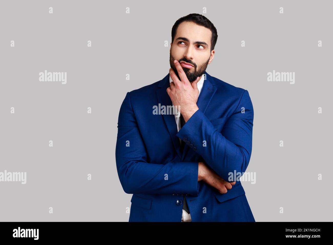 Pensive thoughtful bearded businessman holding his chin with hand, pondering about future, planning strategy, wearing official style suit. Indoor studio shot isolated on gray background. Stock Photo