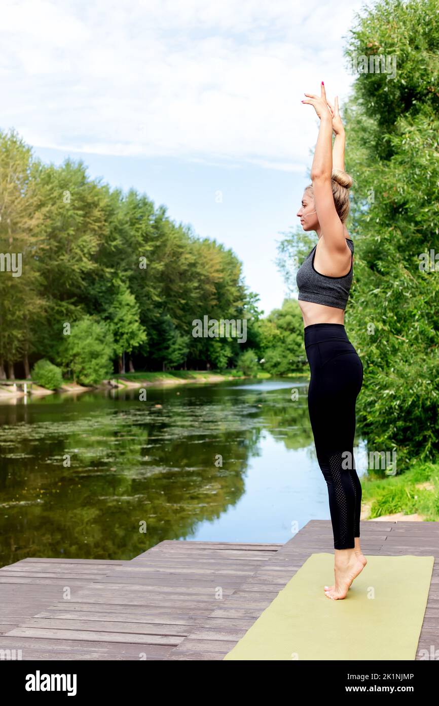 A woman stands on a green rug, by a pond , in summer, doing yoga. Stock Photo