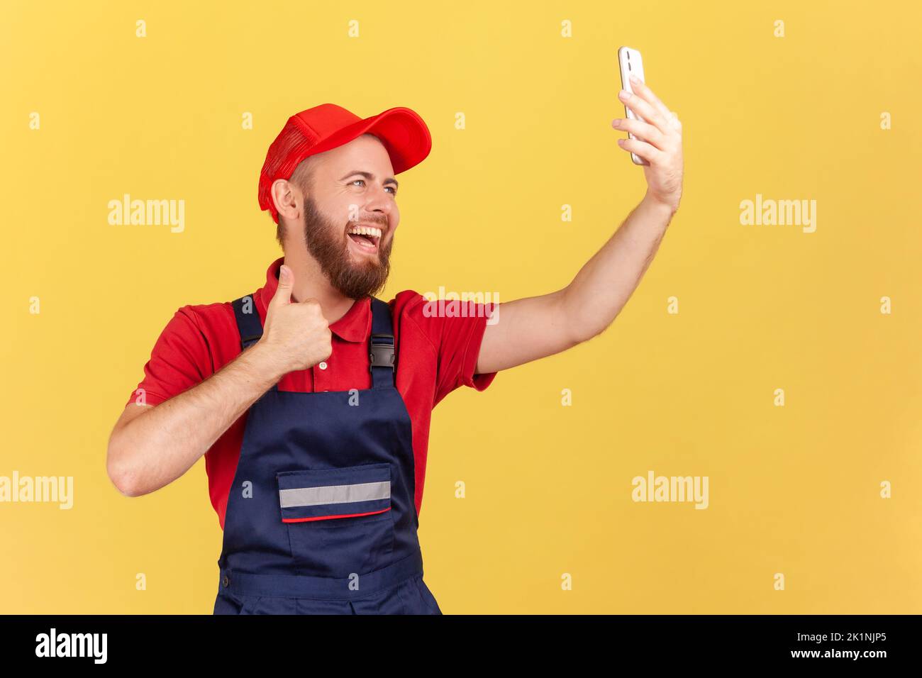 Portrait of positive worker in overalls taking selfie or talking on video call, looking at device camera and showing thumb up to client. Indoor studio shot isolated on yellow background. Stock Photo