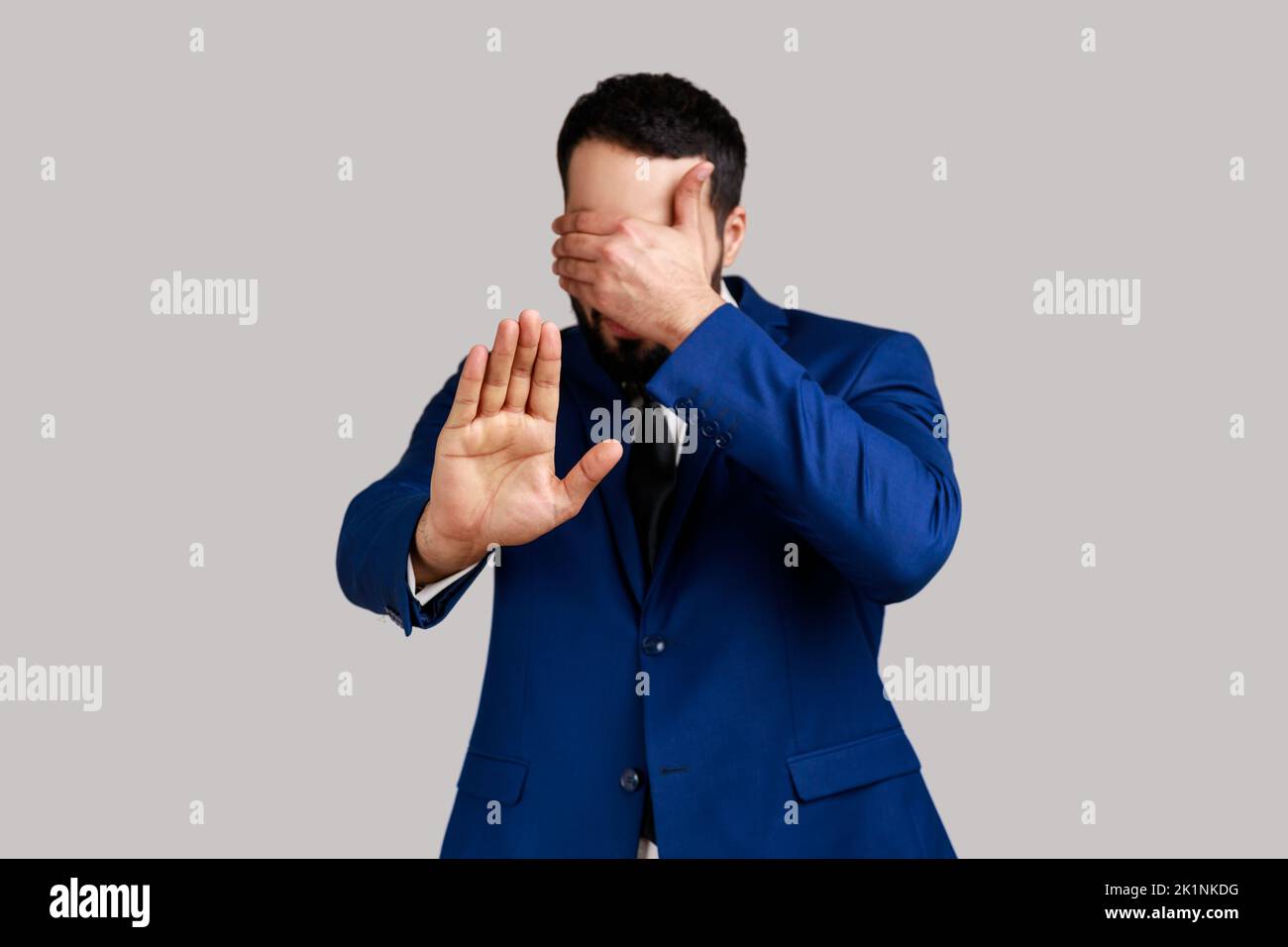Man closing eyes with hand, outstretching palm, dont want to see that, ignoring problems, hiding from stressful situations, wearing official style suit. Indoor studio shot isolated on gray background. Stock Photo