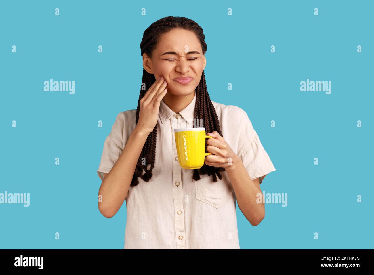 Portrait of unhealthy woman with black dreadlocks wearing white shirt. holding cup in hands, having tooth pain after drinking cold or hot beverage. Indoor studio shot isolated on blue background. Stock Photo