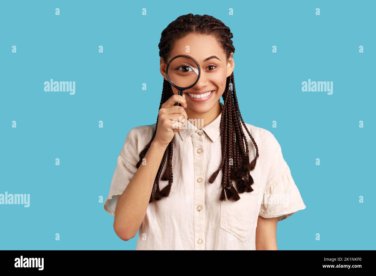 Portrait of funny woman with black dreadlocks holding magnifying glass and looking at camera with big zoom eye, positive face, wearing white shirt. Indoor studio shot isolated on blue background. Stock Photo