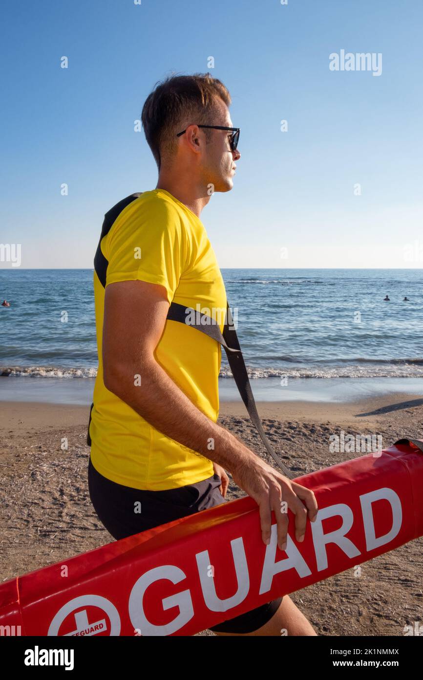 A male lifeguard wiyh yellow shirt on the Mediterranean beach watching people in water. Safety while swimming, handsome brunette male lifeguard. Stock Photo