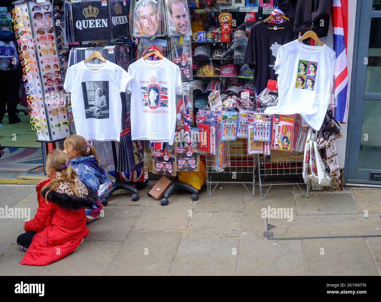 Two children sit outside a shop in Windsor selling royal memorabilia. T-shirts on display. Children with backs to camera. Stock Photo