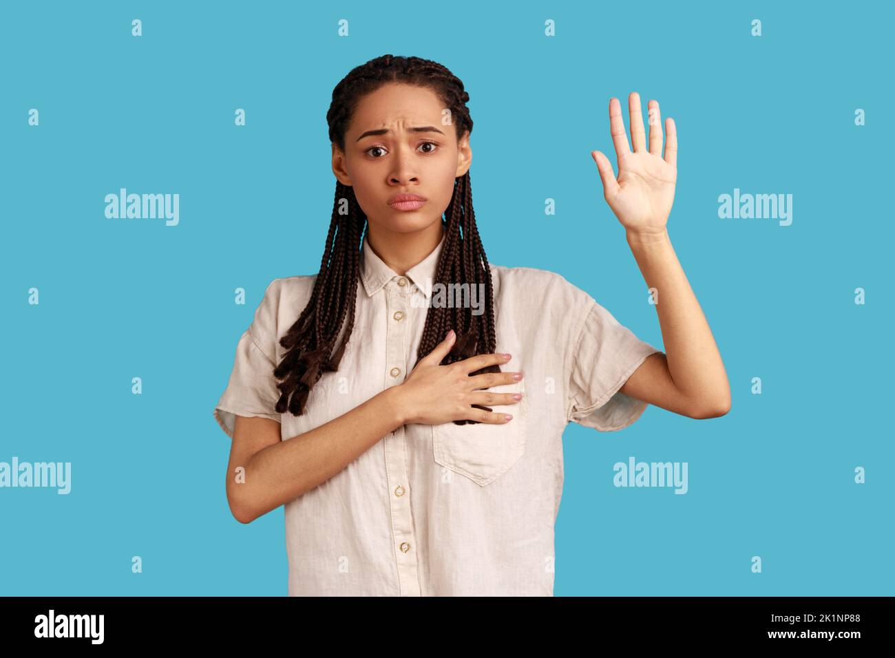 I promise. Portrait of woman with dreadlocks making swearing gesture and holding arm on chest, taking oath, pledging allegiance, wearing white shirt. Indoor studio shot isolated on blue background. Stock Photo