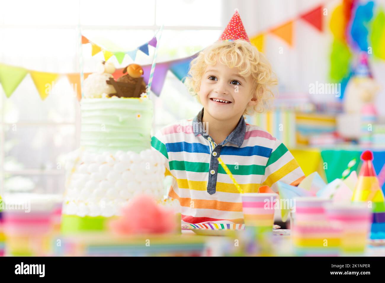 Kids birthday party. Child blowing out candles on colorful cake. Decorated home with rainbow flag banners, balloons. Farm animals theme celebration. L Stock Photo