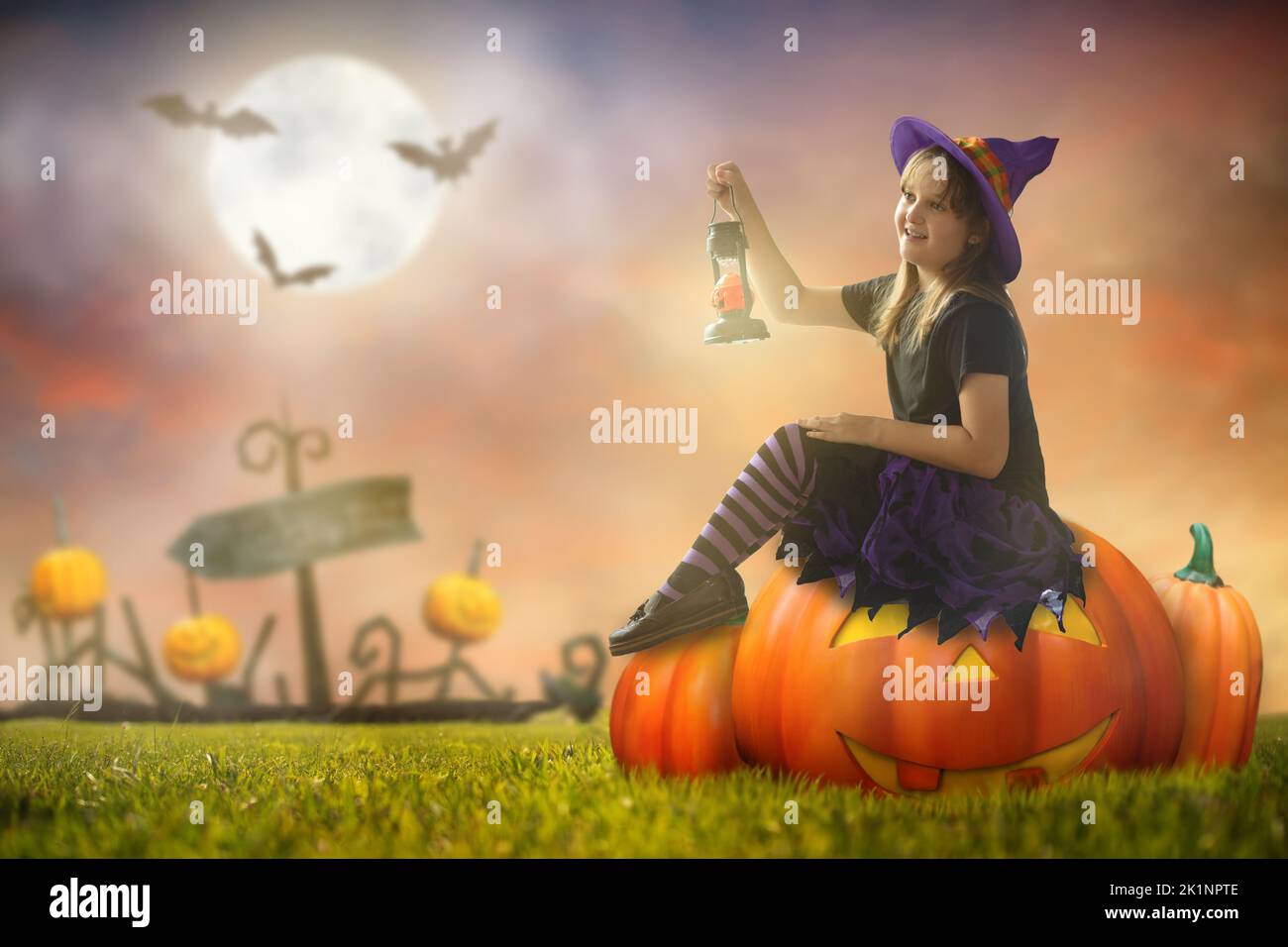 Little witch sitting on giant pumpkin lantern on Halloween night. Huge full moon and bats in the background. Kids trick or treat costume. Children hav Stock Photo