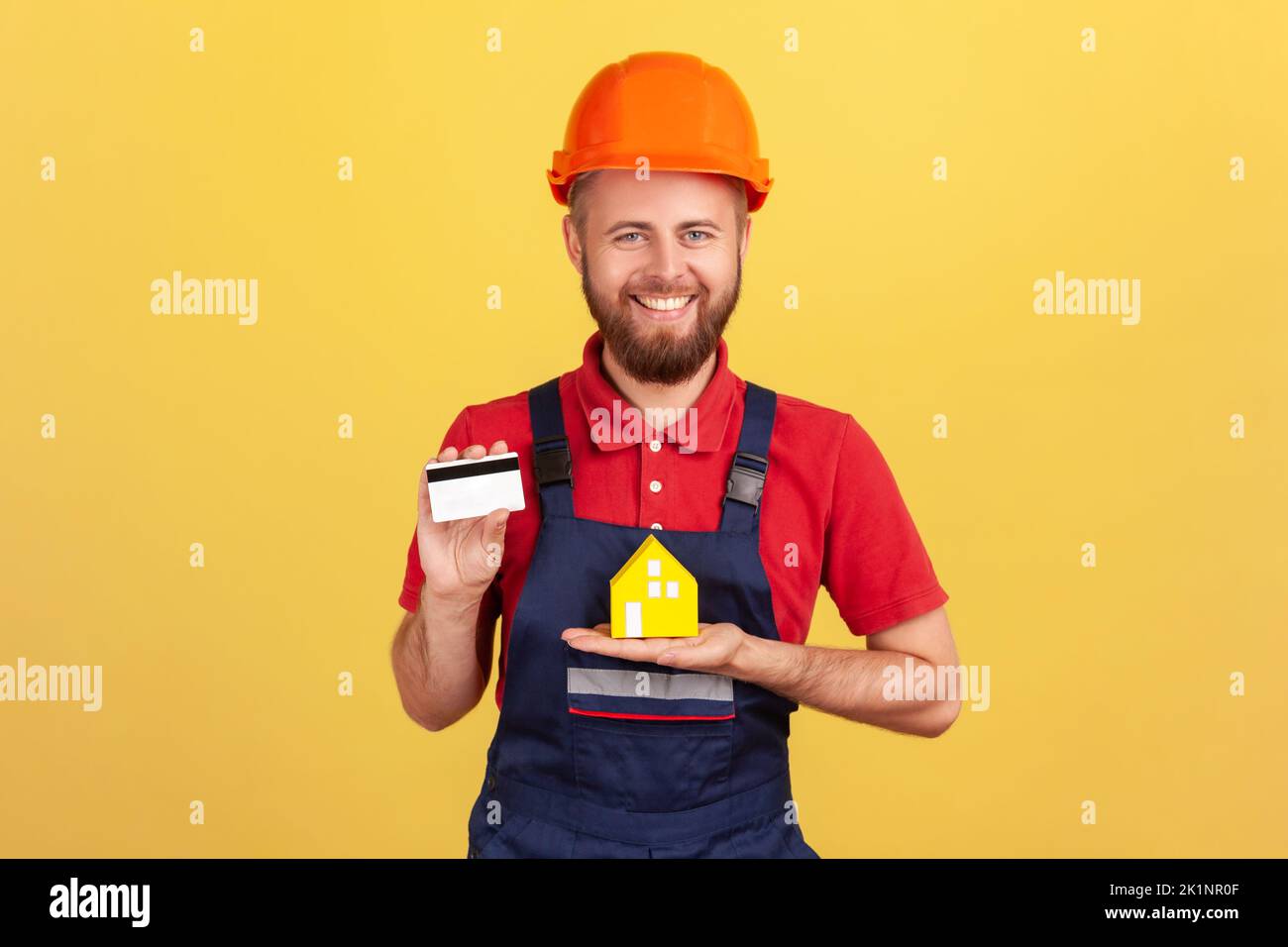 Portrait of happy builder man wearing blue uniform and protective helmet showing credit card and paper house, looking at camera with smile. Indoor studio shot isolated on yellow background. Stock Photo