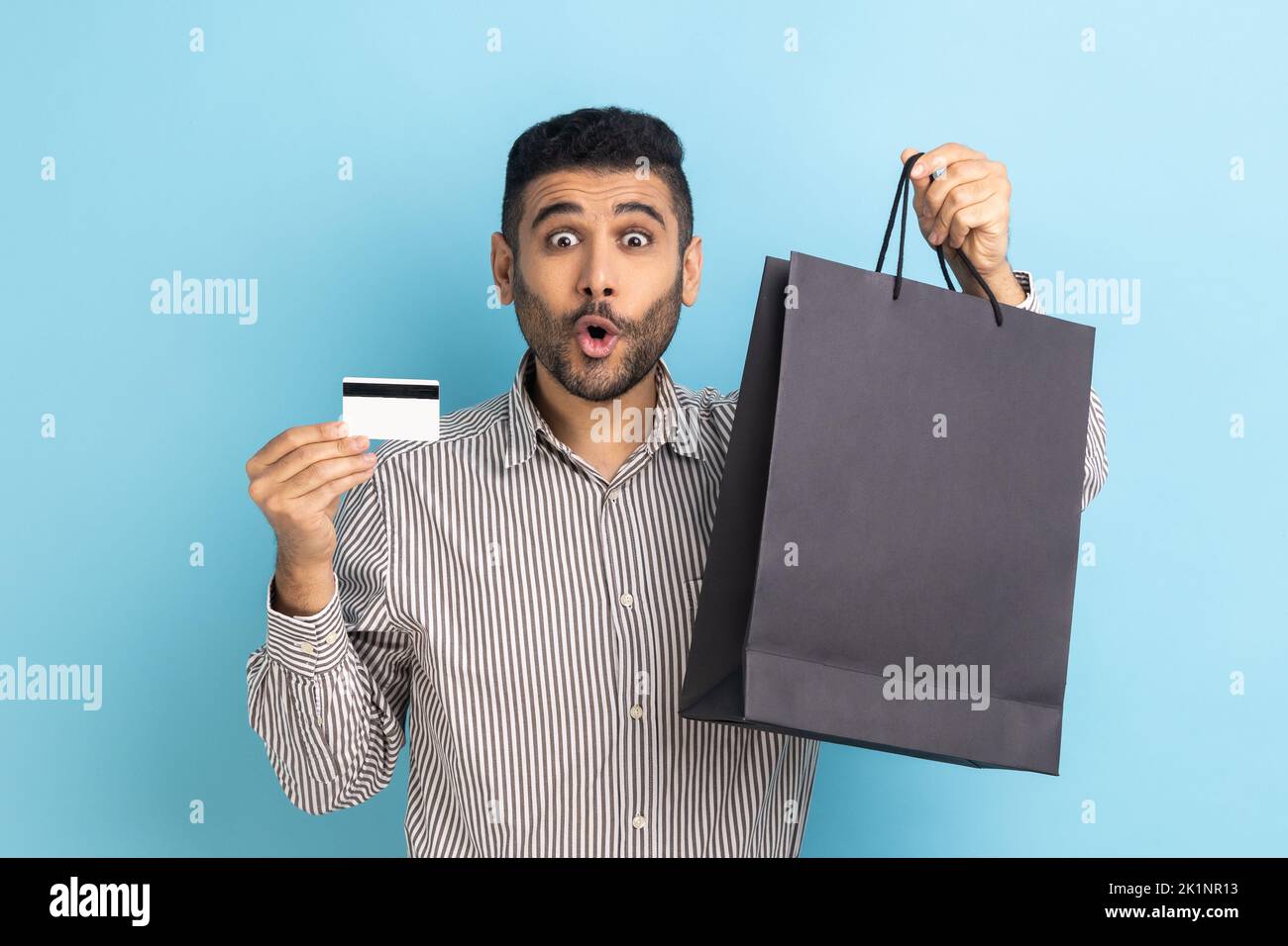 Astonished businessman holding and showing limitless credit card and shopping bags, surprised with great shopping, wearing striped shirt. Indoor studio shot isolated on blue background. Stock Photo