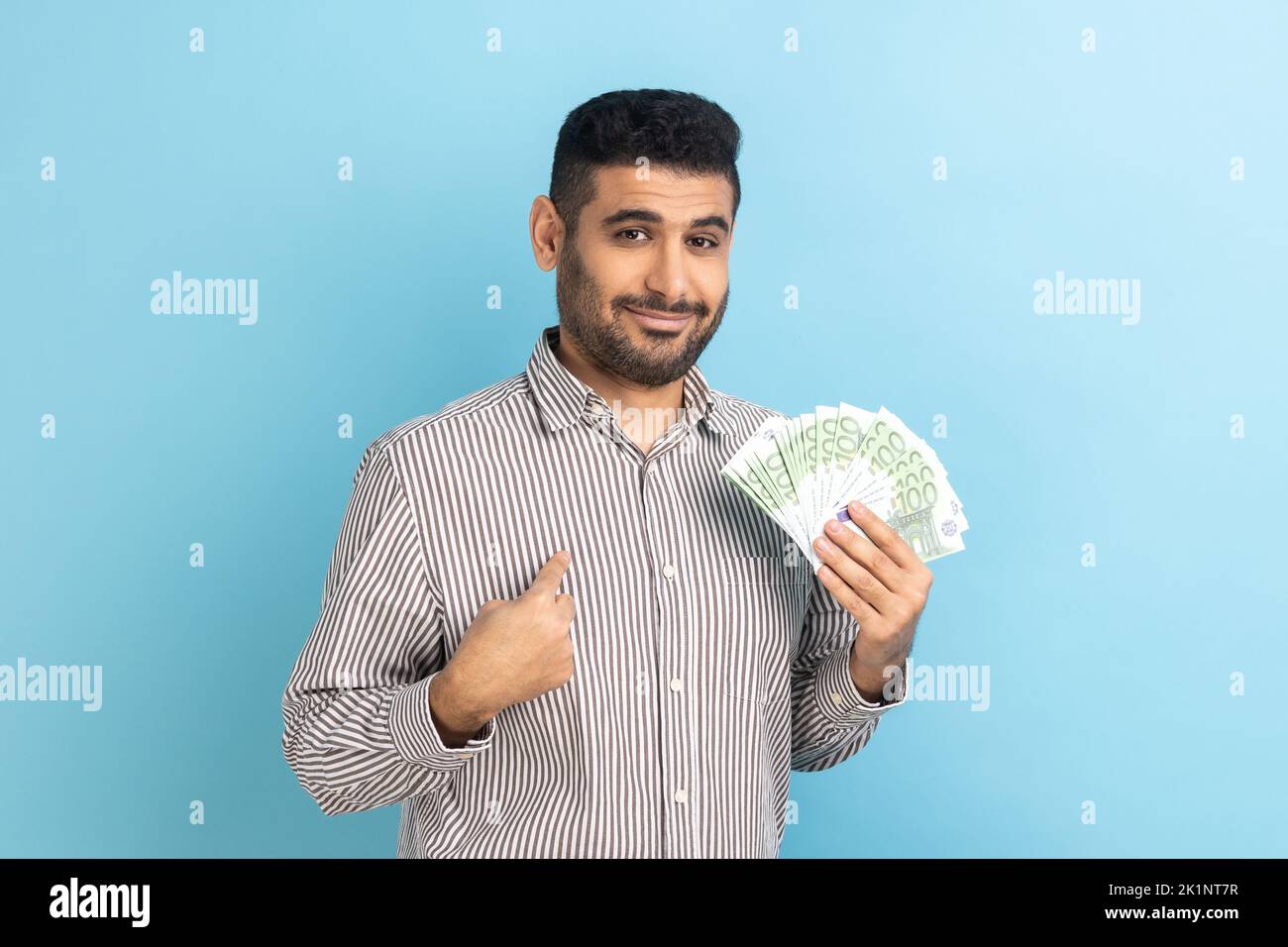 Portrait of young adult businessman with beard holding euro bills and pointing at himself with pride, looking at camera, wearing striped shirt. Indoor studio shot isolated on blue background. Stock Photo