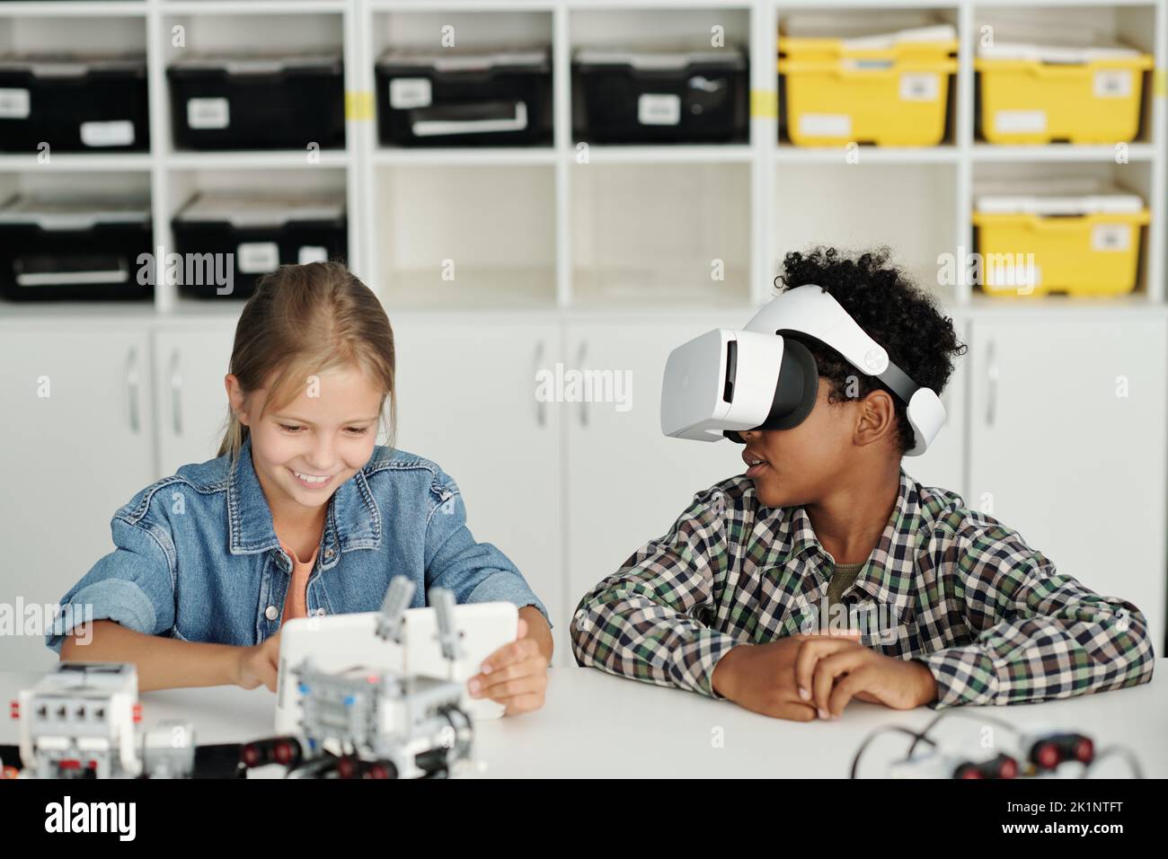 Two clever intercultural schoolkids playing virtual game at lesson of robotics while schoolgirl adjusting settings in tablet Stock Photo
