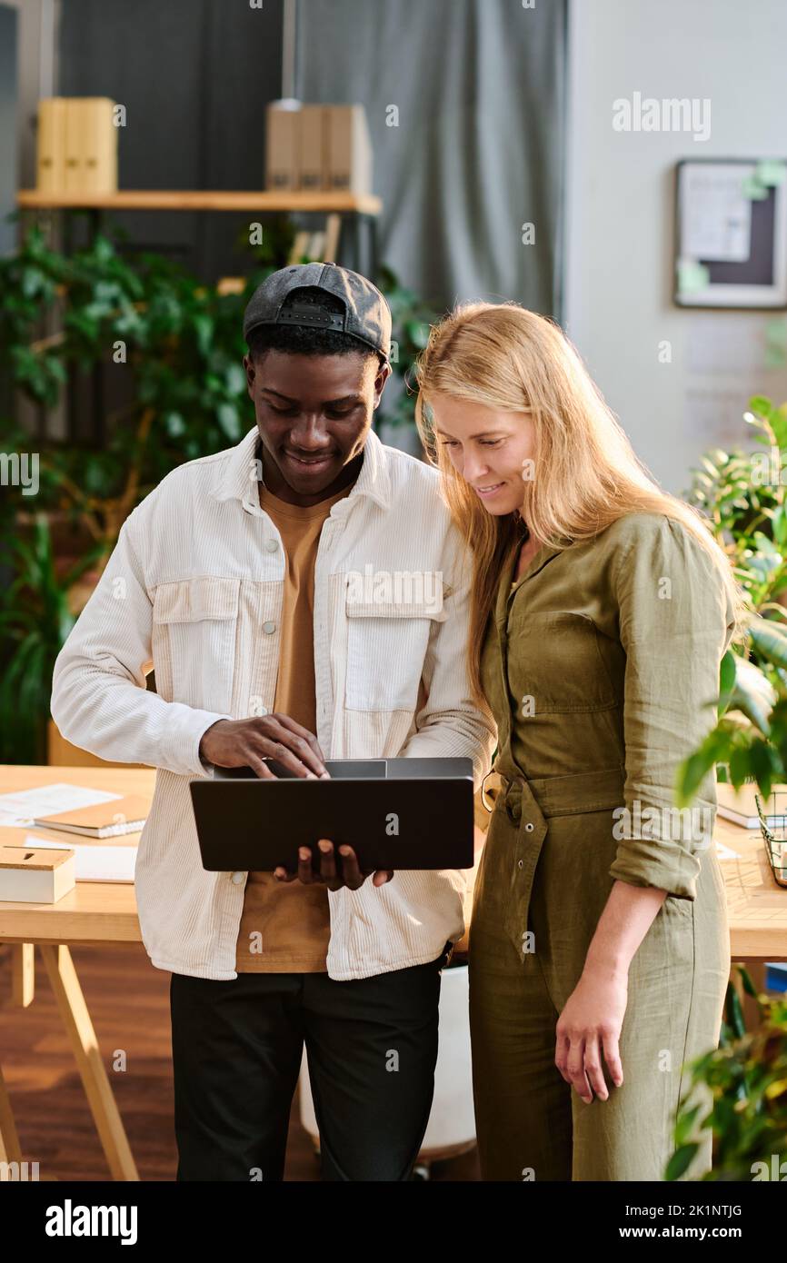 Two young intercultural employees in casualwear looking at laptop screen while standing by desk and preparing presentation of project Stock Photo