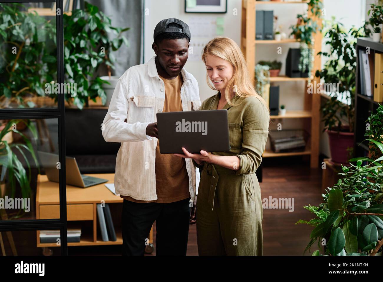 Two young intercultural employees in casualwear looking at screen of laptop held by blond businesswoman during discussion Stock Photo