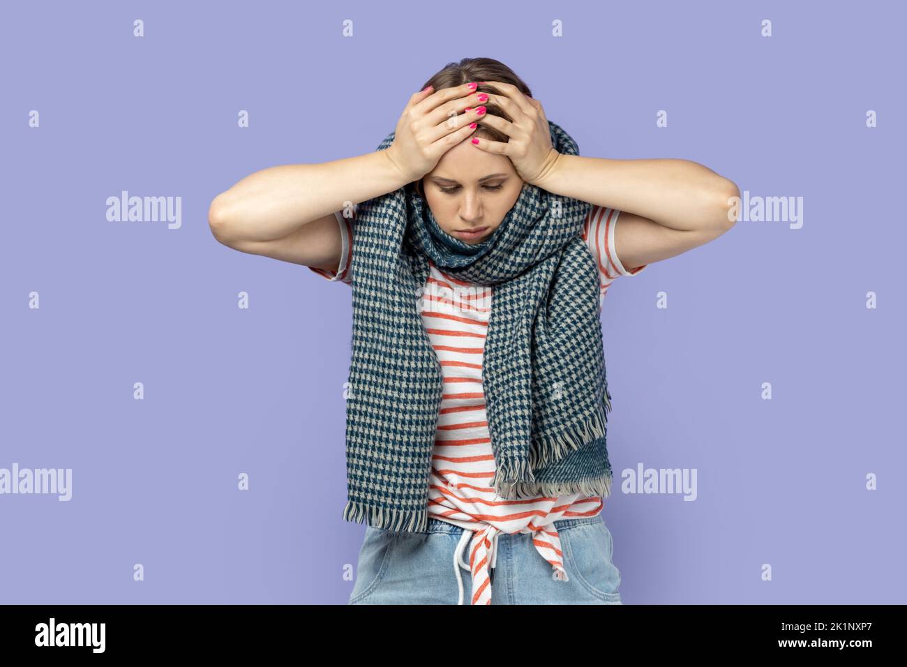 Portrait of sick unhealthy woman wearing striped T-shirt feels unwell, suffering terrible headache, standing wrapped in warm scarf, having influenza. Indoor studio shot isolated on purple background. Stock Photo