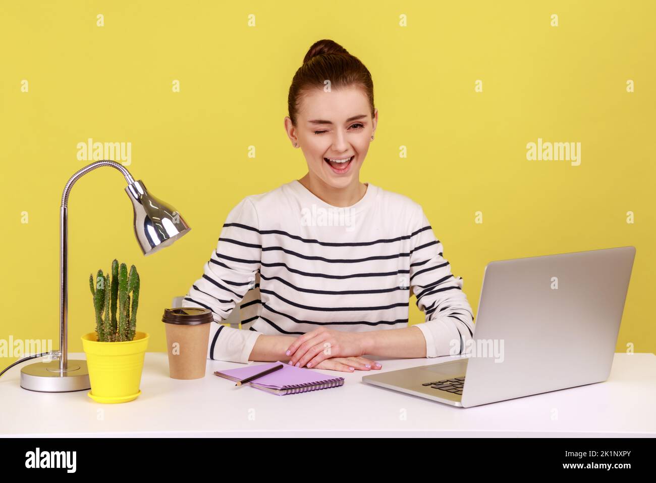 Optimistic woman office worker in striped shirt sitting at workplace and winking, looking at camera with toothy smile, good mood. Indoor studio studio shot isolated on yellow background. Stock Photo