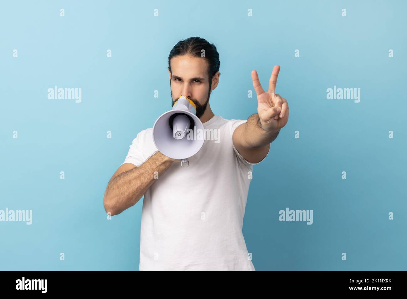 Portrait of handsome man with beard wearing white T-shirt holding megaphone in hands and making announcement, showing v sign to camera. Indoor studio shot isolated on blue background. Stock Photo