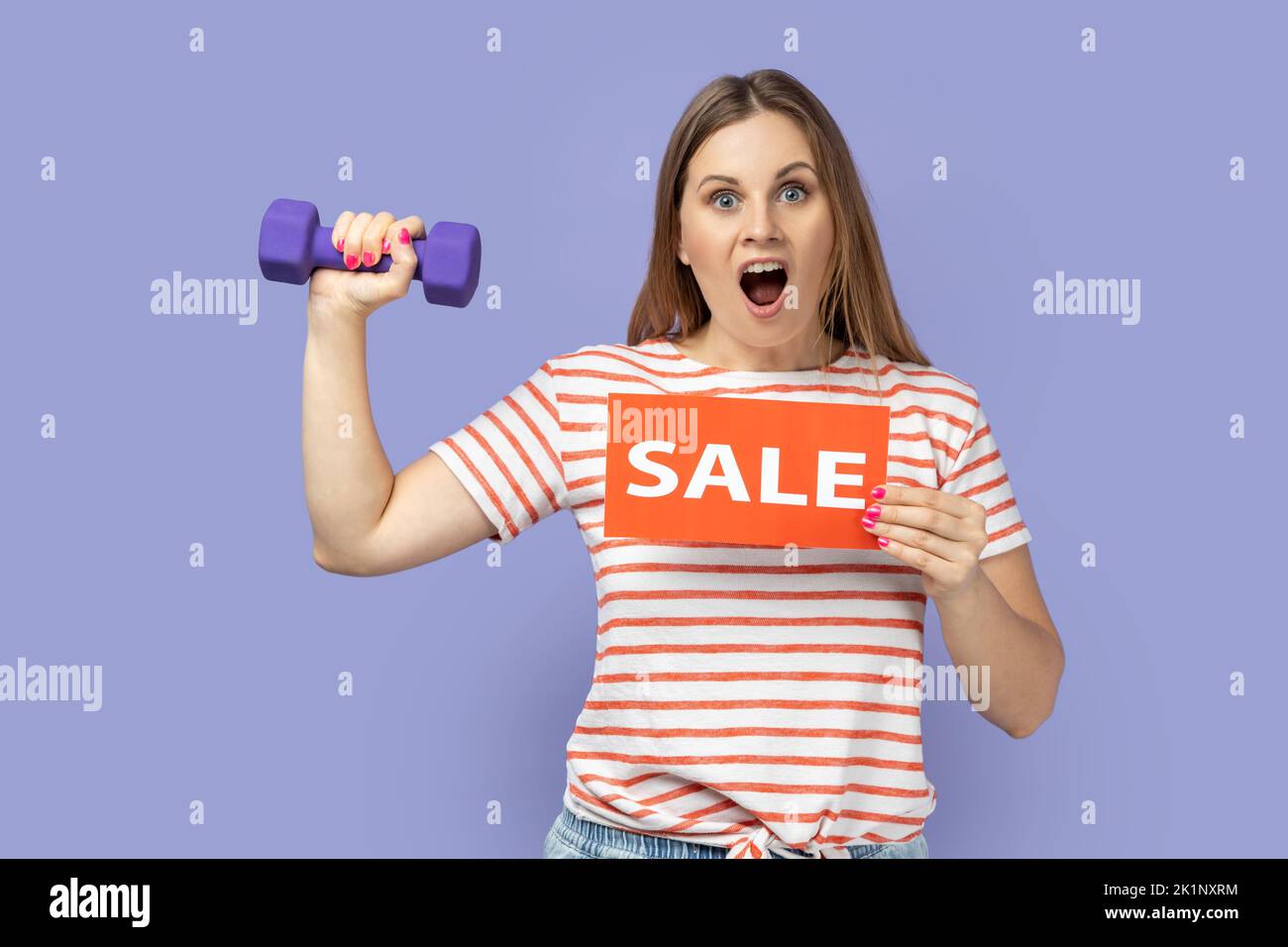 Portrait of amazed excited blond woman wearing striped T-shirt holding card with sale inscription and raised dumbbell, discounts for fitness workout. Indoor studio shot isolated on purple background. Stock Photo