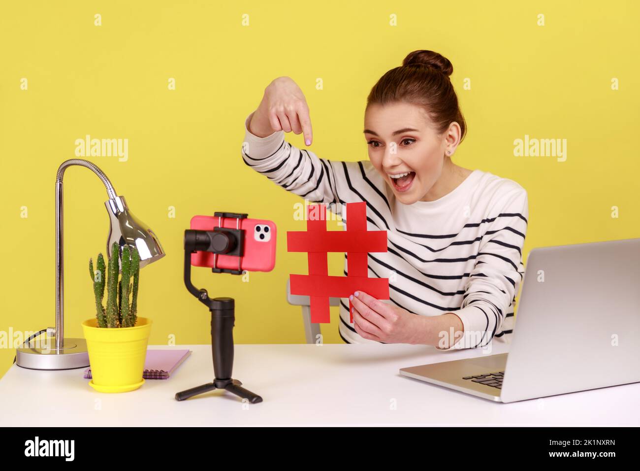Smiling excited woman blogger showing big red hashtag symbol at smartphone camera, sharing viral content, tagged message. Indoor studio studio shot isolated on yellow background. Stock Photo