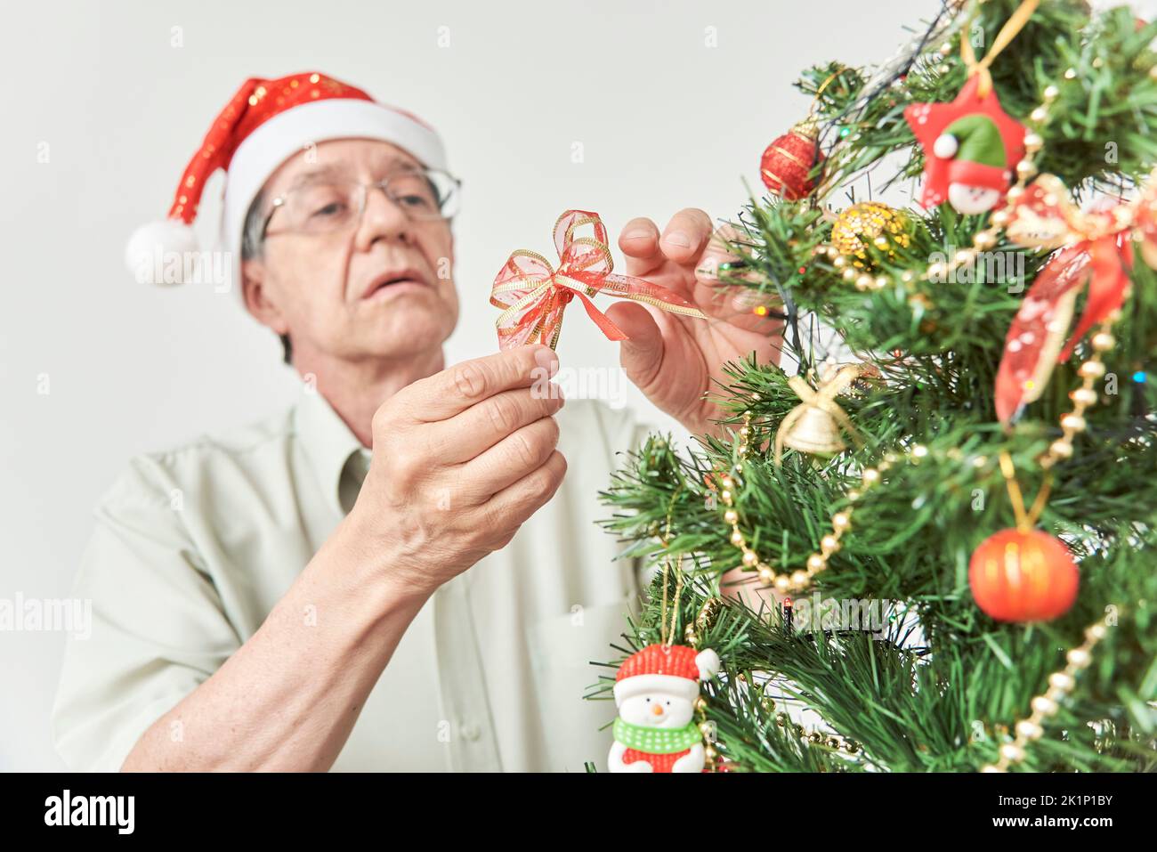 Senior hispanic man decorating, placing a red ribbon bow on a Christmas tree at home wearing a red Santa Claus beanie. Stock Photo
