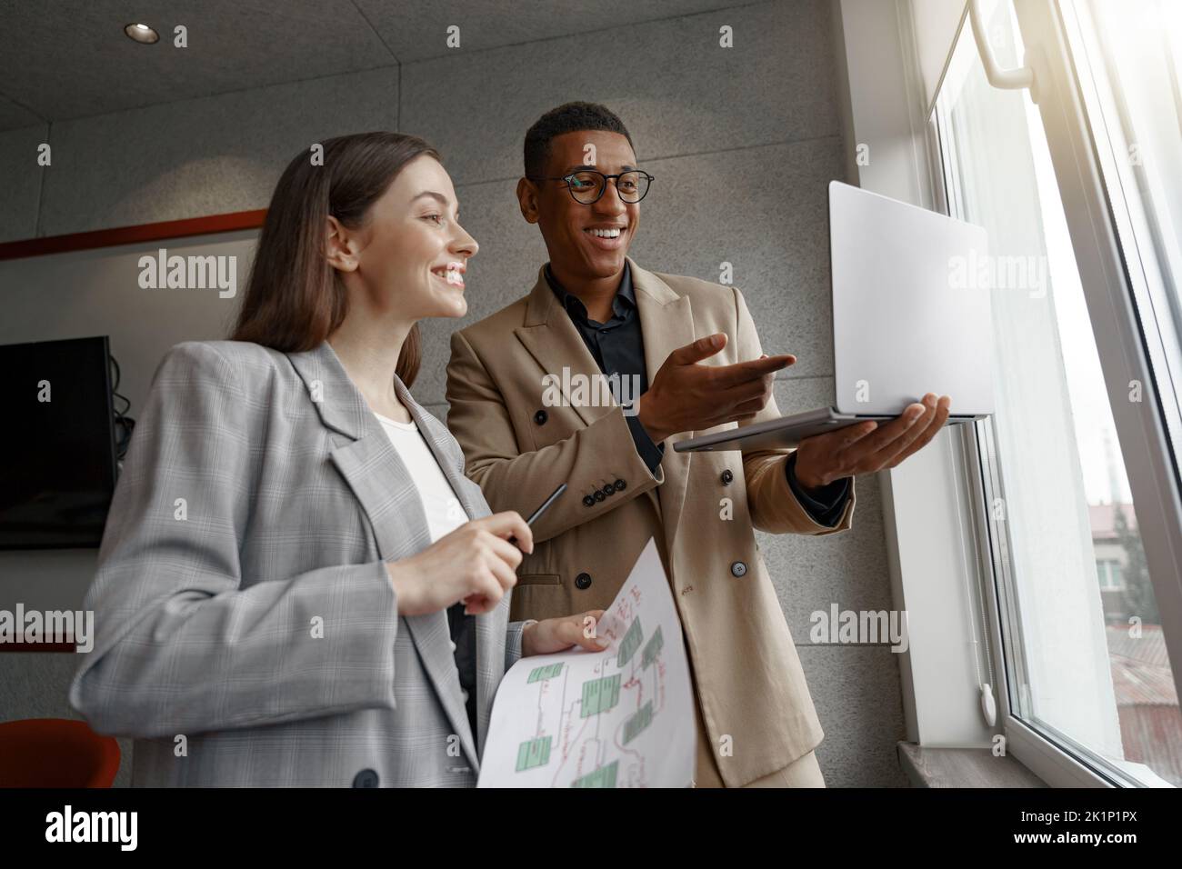 Two smiling business people working with laptop together standing near window Stock Photo