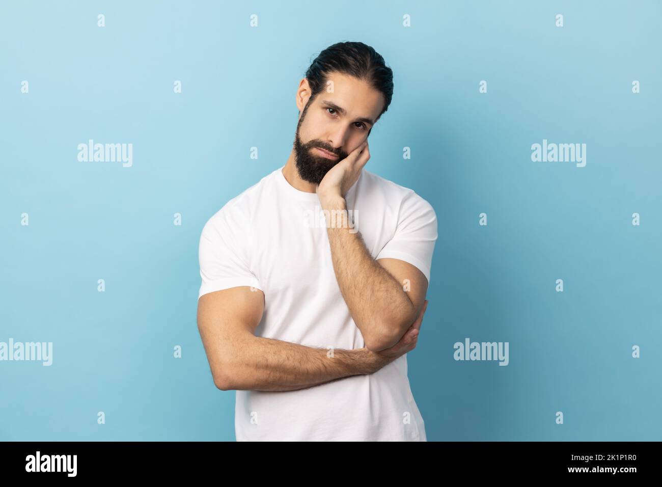 Portrait of man with beard wearing white T-shirt leaning head on hand looking at camera with indifferent look, laziness and apathy, procrastination. Indoor studio shot isolated on blue background. Stock Photo