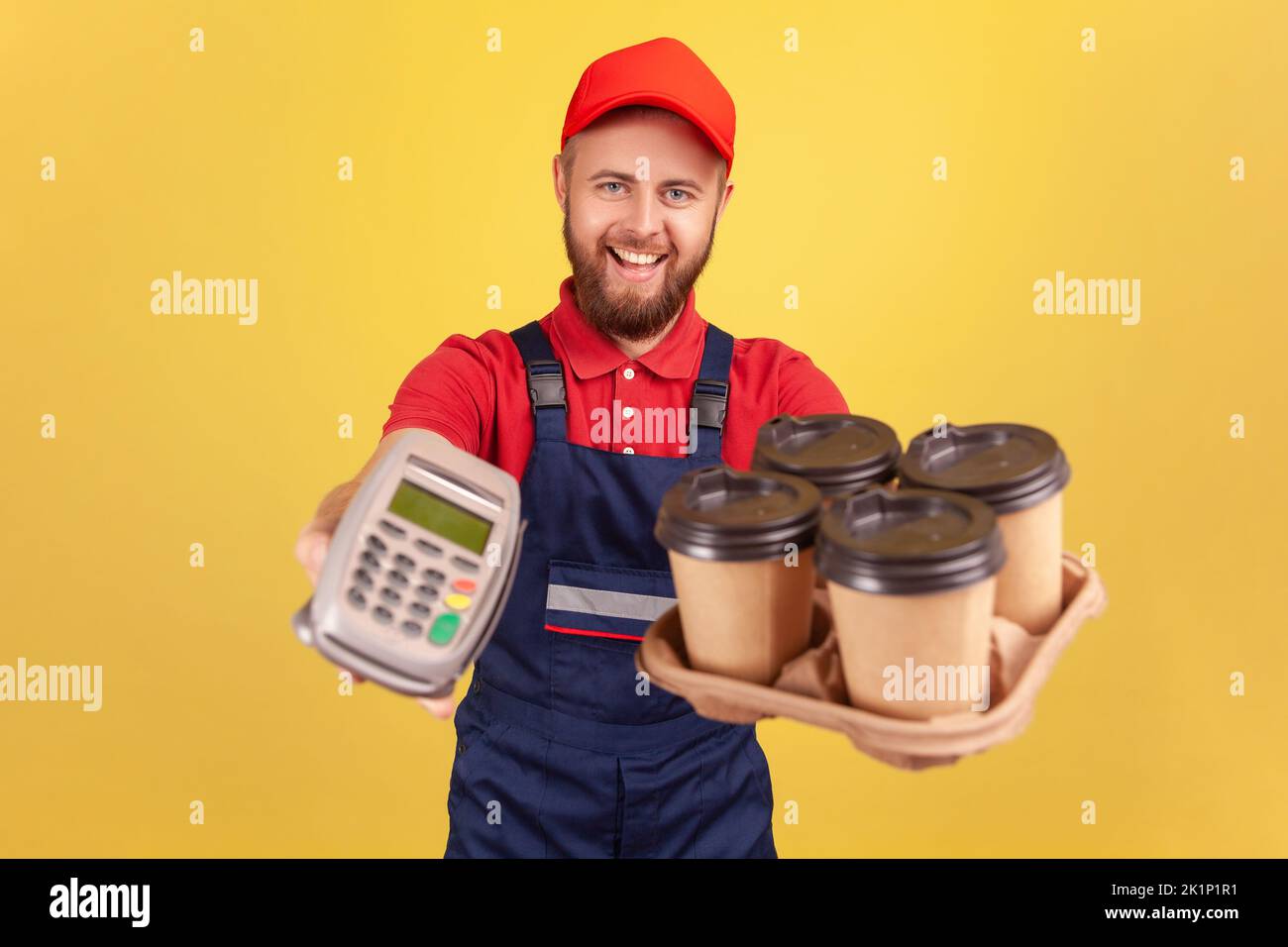 Friendly optimistic delivery man holding terminal and coffee with pizza box, looking at camera with happy expression, wearing overalls and cap. Indoor studio shot isolated on yellow background. Stock Photo