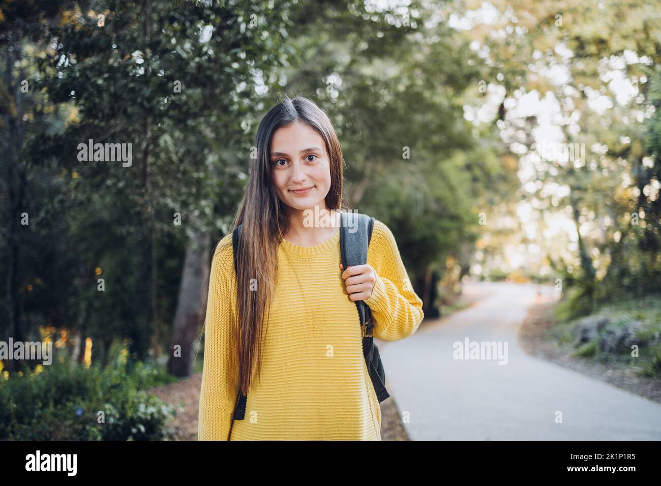 Smiling female college student wearing a yellow sweater and a backpack in the campus park road. Innocent smile Stock Photo