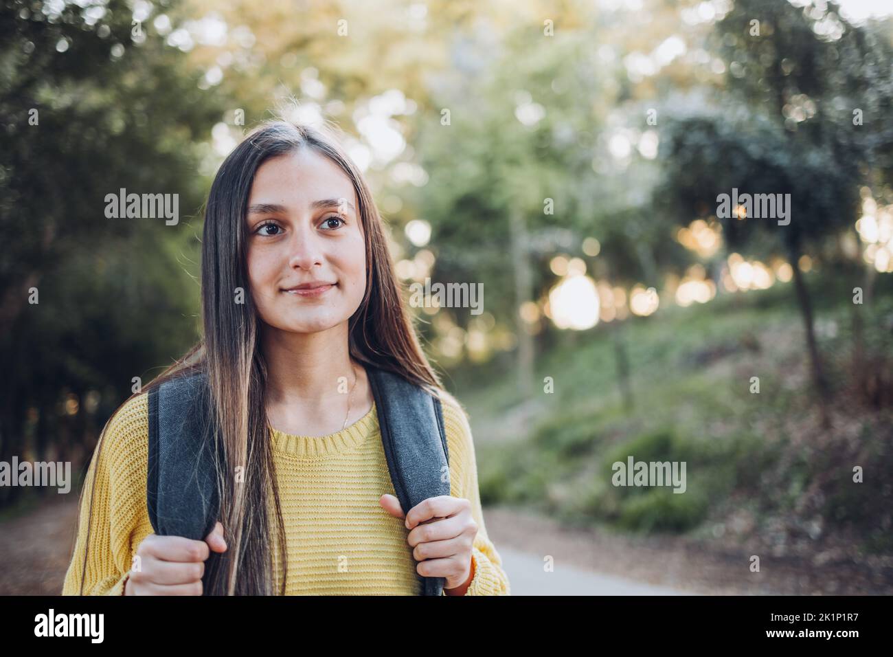 Smiling female university student wearing a yellow sweater and a backpack in the campus park road. Innocent smile Stock Photo
