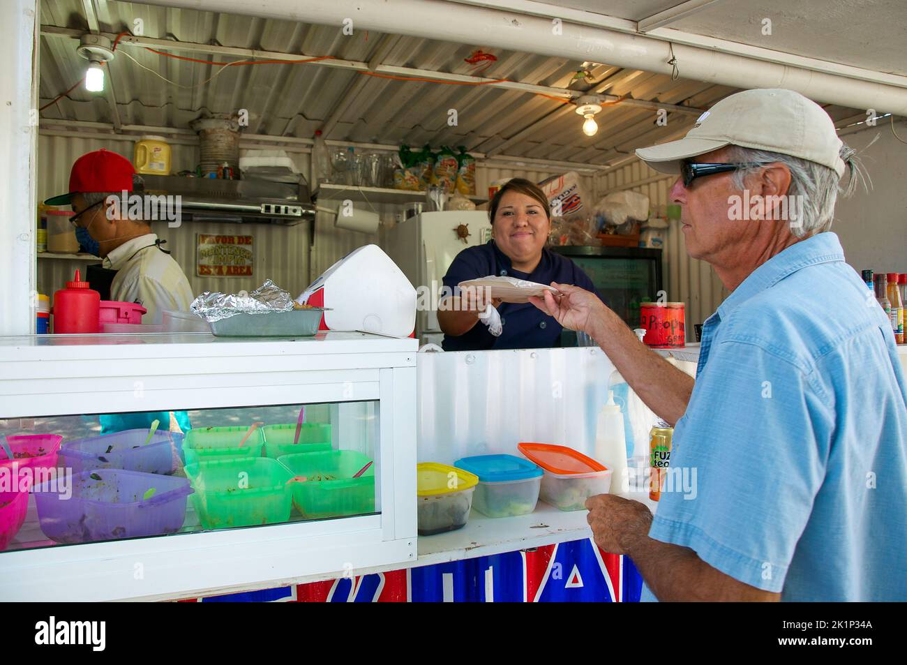 A smiling woman working in a taco stand hands a paper plate with food to a senior male, Baja Sur, Mexico. Stock Photo