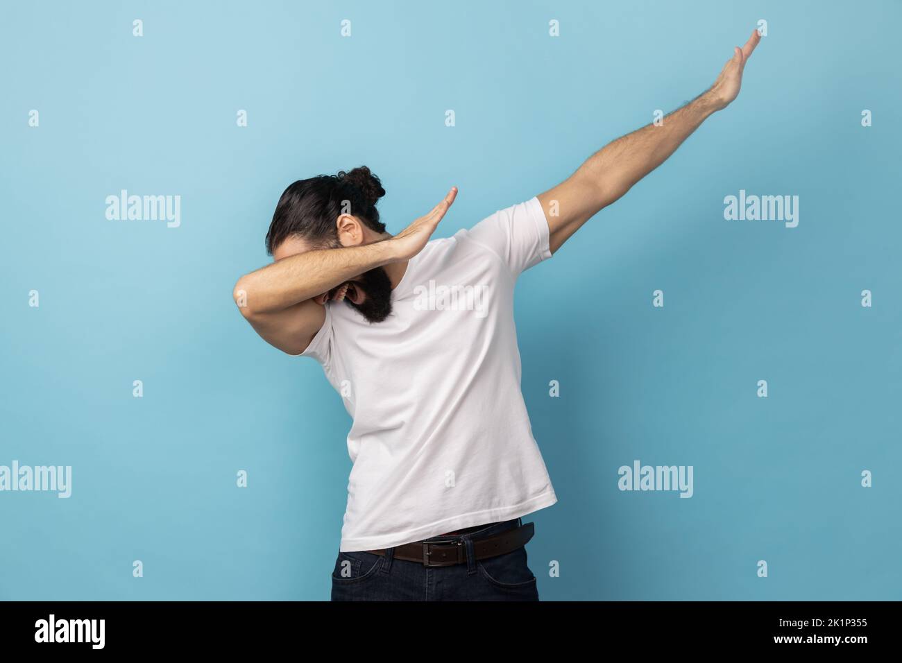Portrait of anonymous unknown man with beard wearing white T-shirt standing in dab dance pose, internet meme, celebrating success. Indoor studio shot isolated on blue background. Stock Photo