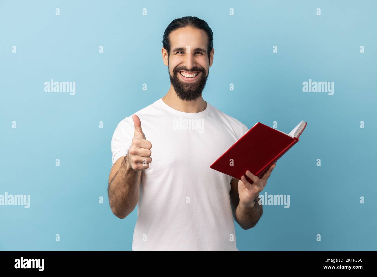 Portrait of positive happy man with beard wearing white T-shirt showing thumbs up gesture holding and reading book, likes genre and plot. Indoor studio shot isolated on blue background. Stock Photo