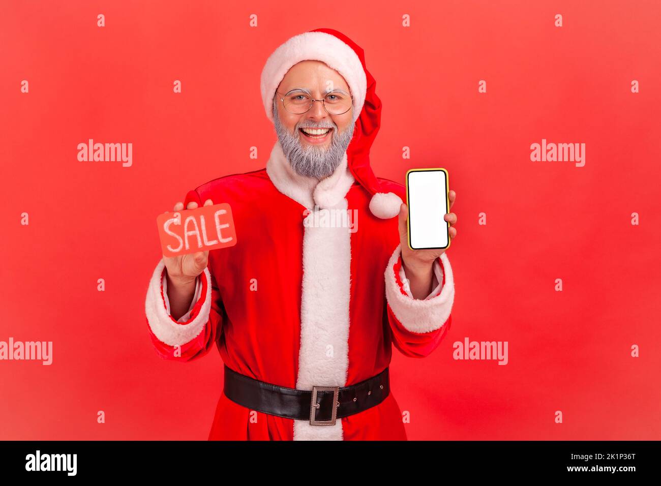 Elderly man with gray beard wearing santa claus costume holding smartphone with blank display and sale card in hands, discount application. Indoor studio shot isolated on red background. Stock Photo