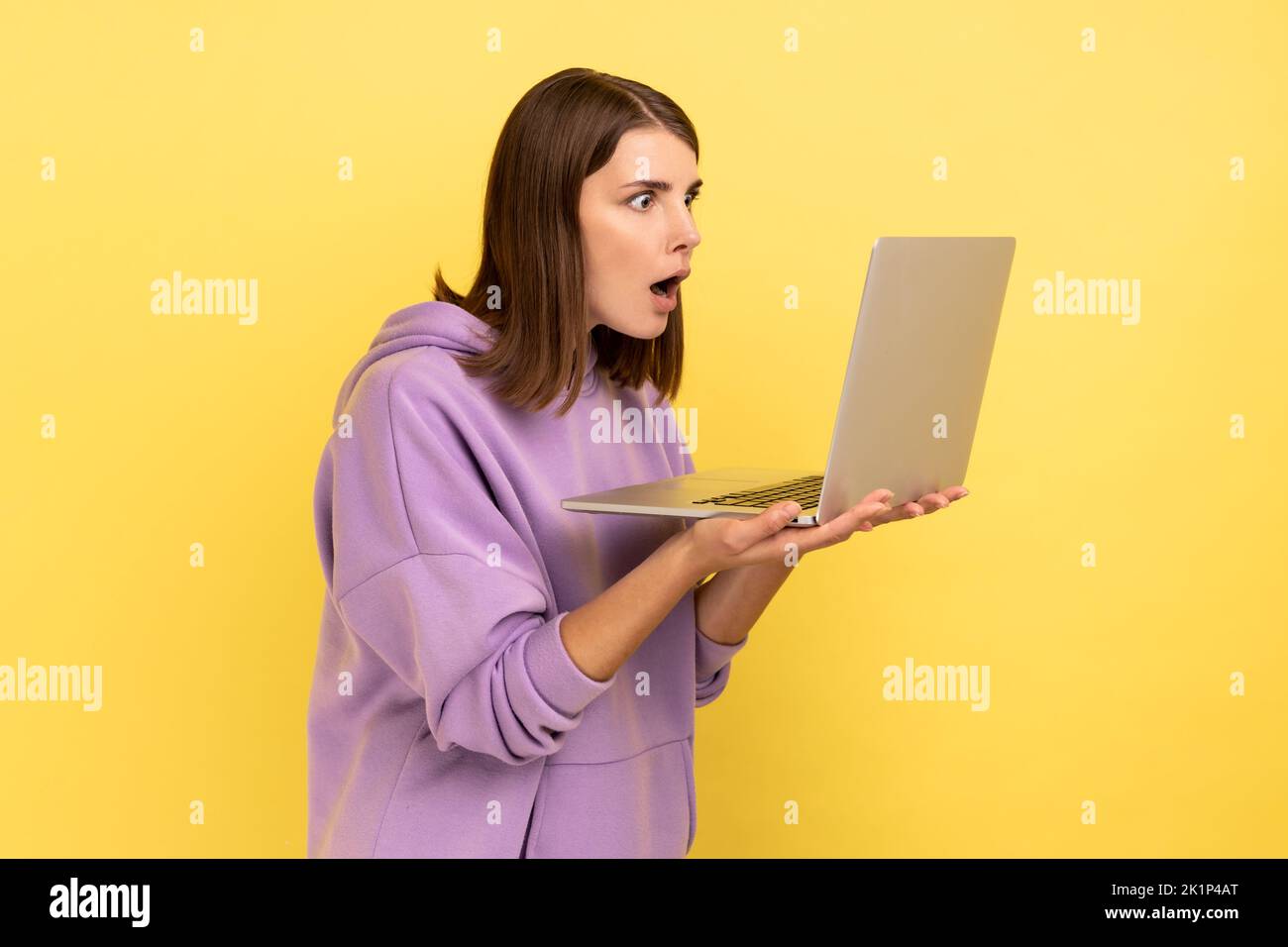 Portrait of amazed shocked young adult woman working on laptop compute, looking at display with open mouth, wearing purple hoodie. Indoor studio shot isolated on yellow background. Stock Photo