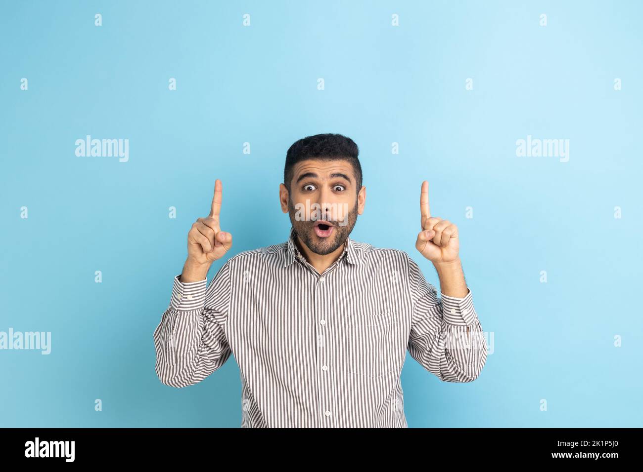 Wow, look, crazy advertisement. Bearded businessman pointing up at empty place for ad content and expressing astonishment, wearing striped shirt. Indoor studio shot isolated on blue background. Stock Photo
