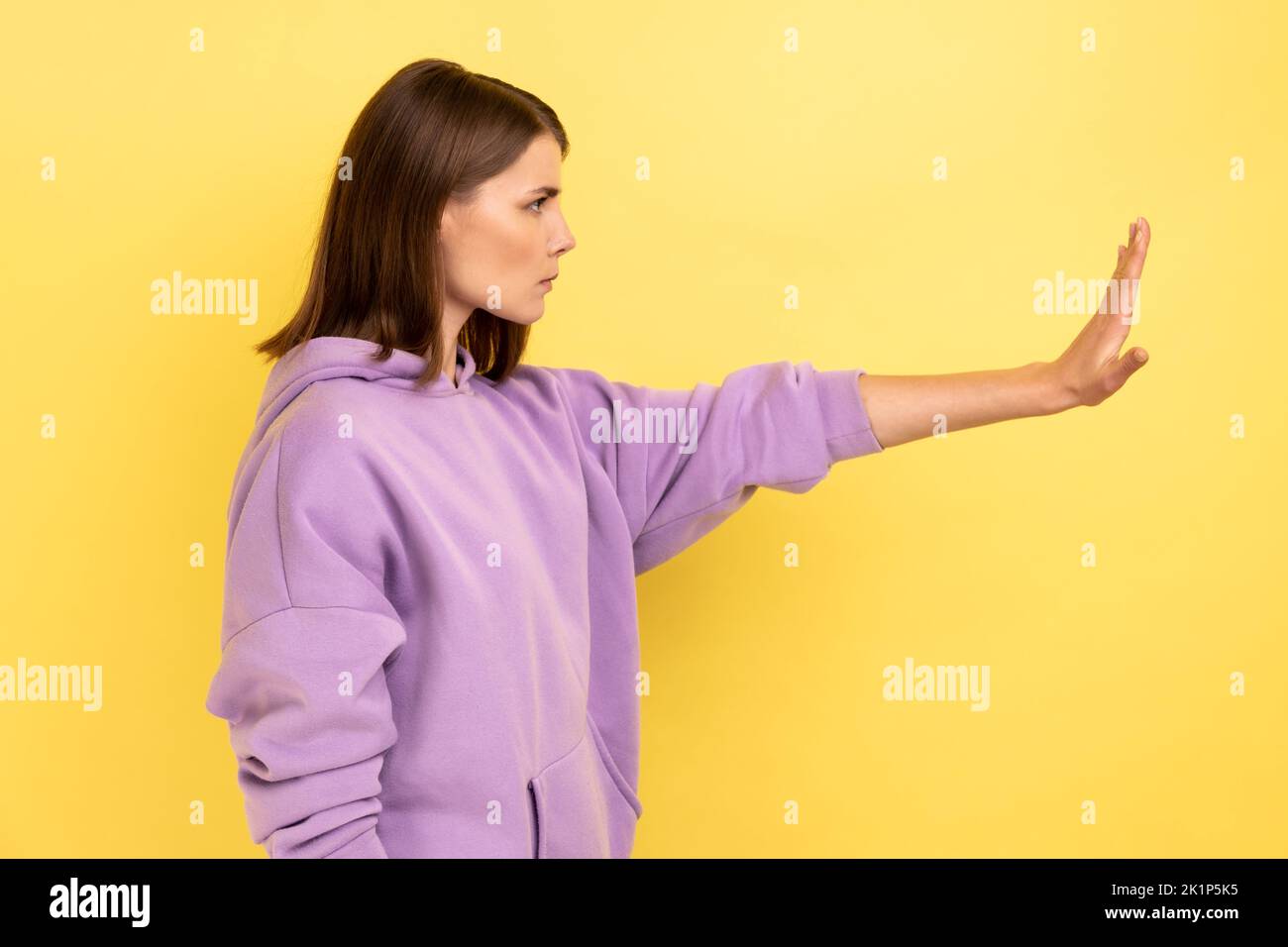 Side view of confused woman showing stop gesture with palm of hand, trying to stop abuser, rejection, afraid of domestic violence, wearing hoodie. Indoor studio shot isolated on yellow background Stock Photo