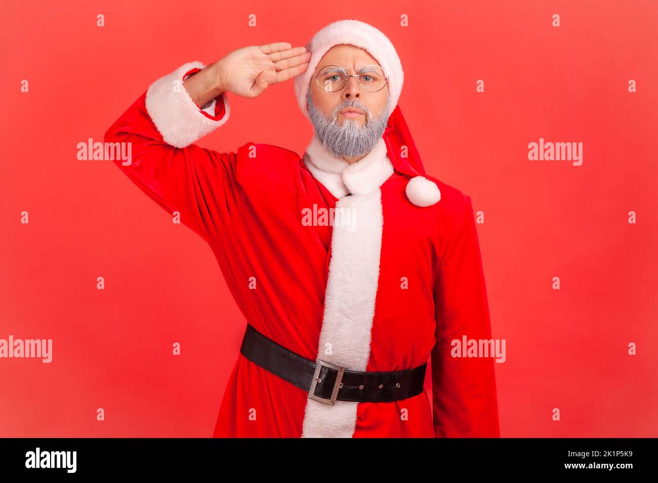 Yes, sir. Portrait of serious obedient elderly man wearing santa claus costume saluting with arm near temple and listening attentively to order. Indoor studio shot isolated on red background. Stock Photo