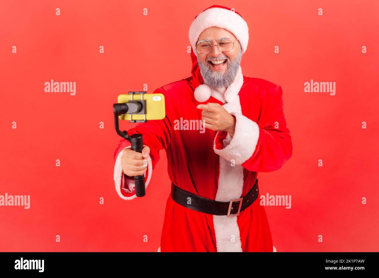 Elderly blogger man with gray beard wearing santa claus costume broadcasting livestream, using phone and steadicam, pointing finger to device display. Indoor studio shot isolated on red background. Stock Photo