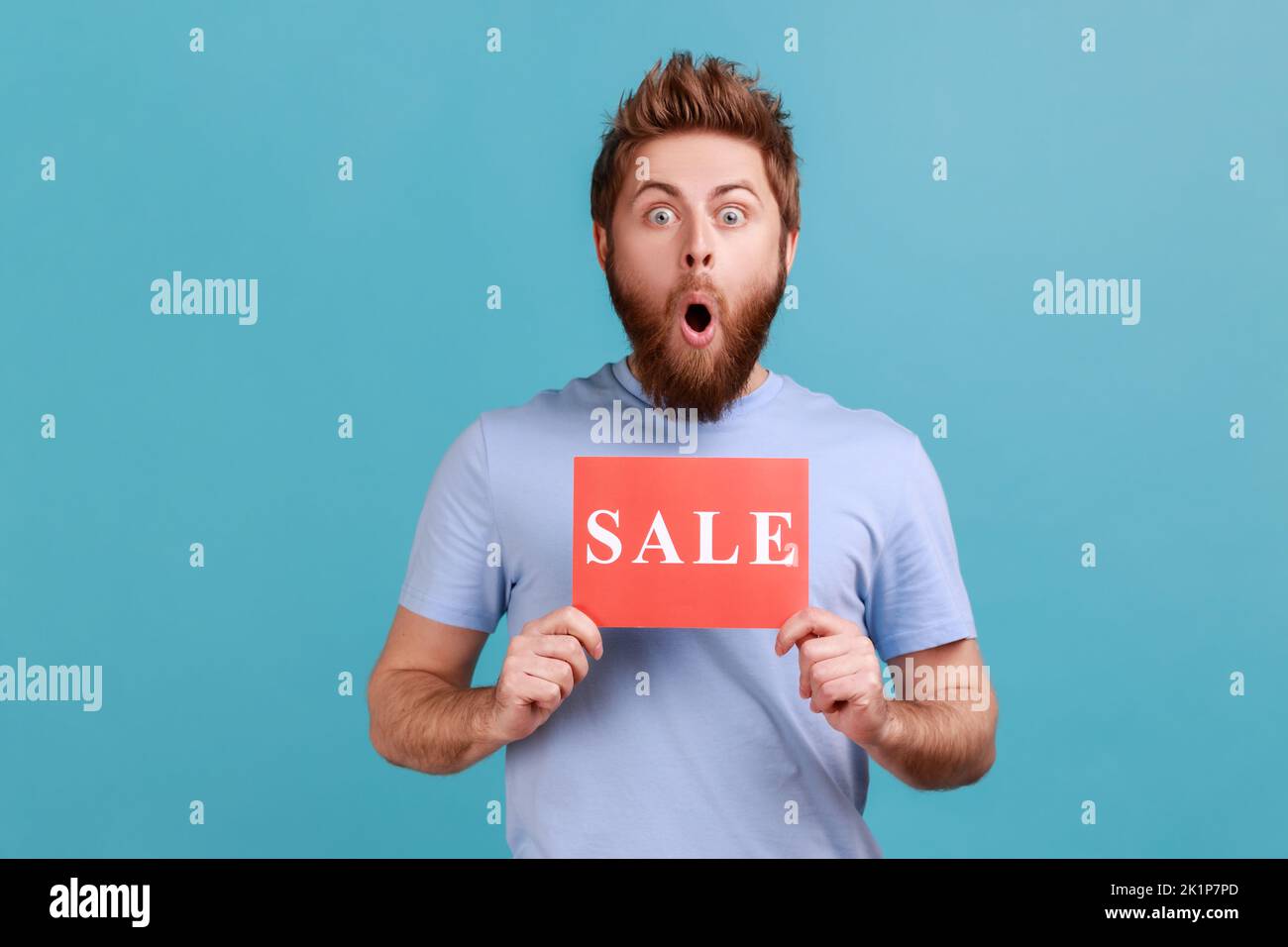 Portrait of surprised amazed bearded man holding card with sale inscription, standing with open mouth and big eyes, astonishing discounts. Indoor studio shot isolated on blue background. Stock Photo