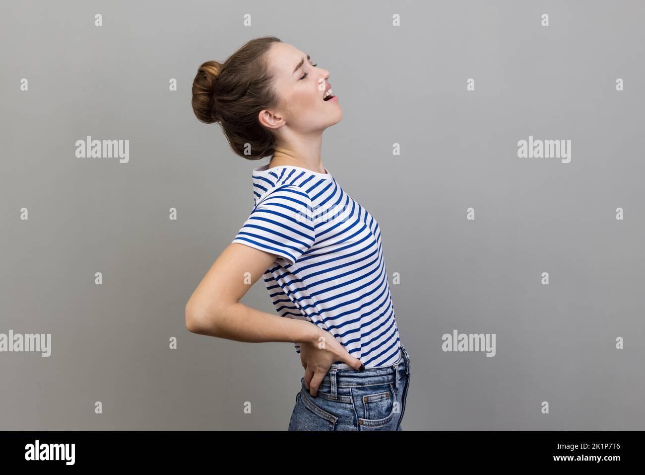 Back, kidney or spine pain. Portrait of woman wearing striped T-shirt standing and endure pain on back, frowning face from pain. Indoor studio shot isolated on gray background. Stock Photo