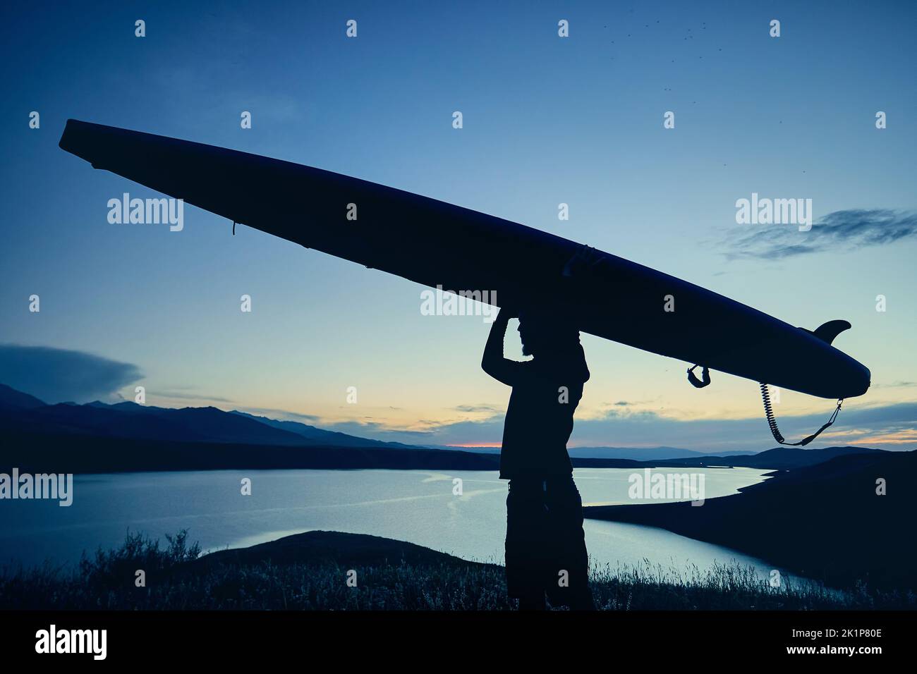 Bearded Man in silhouette holding SUP board near lake at sunset in Kazakhstan. Stand up paddle boarding outdoor active recreation in nature. Stock Photo