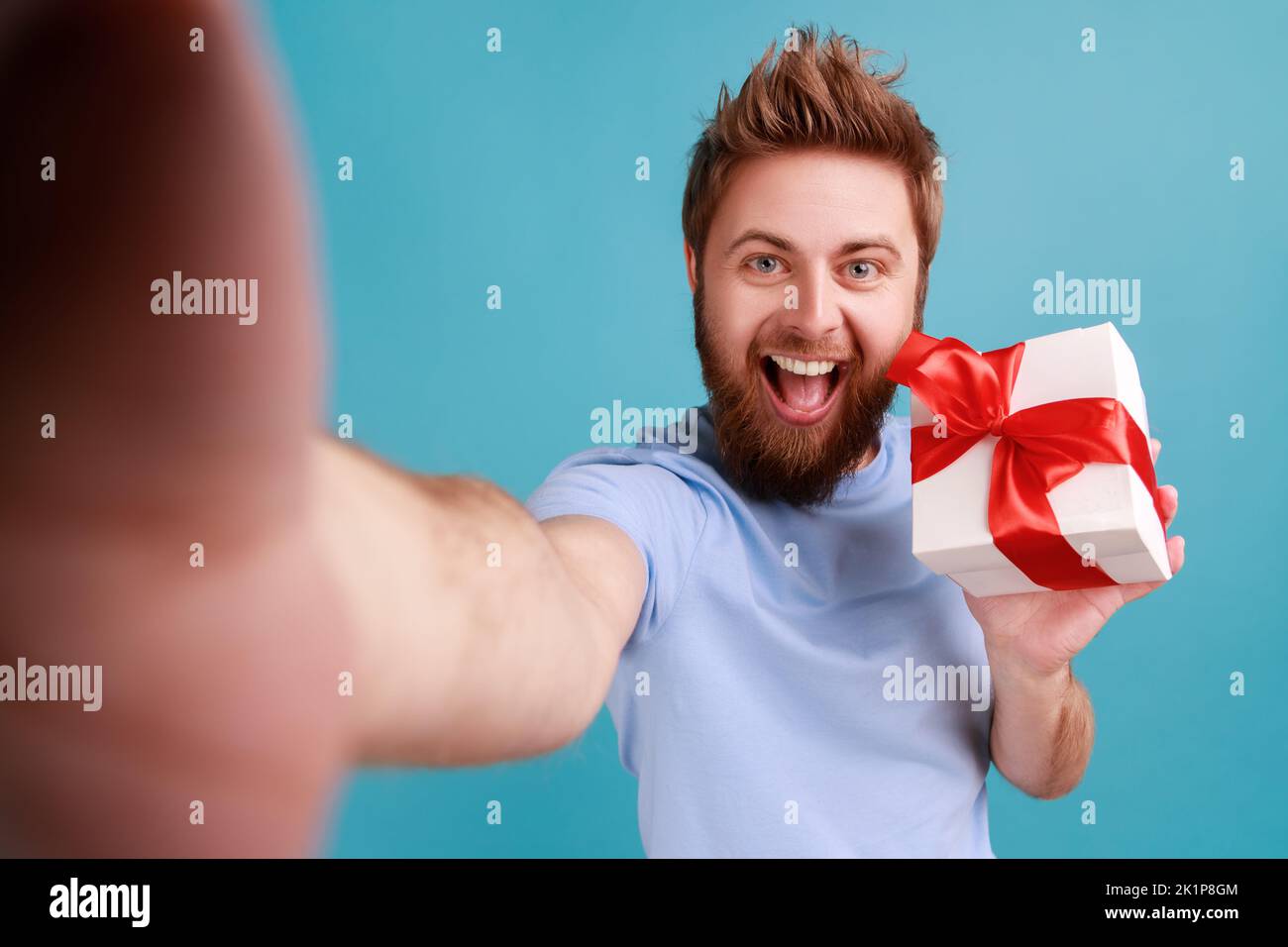 Portrait of positive bearded man with widely opened mouth looking at camera with happy smile POV, point of view of photo, holding present box. Indoor studio shot isolated on blue background. Stock Photo