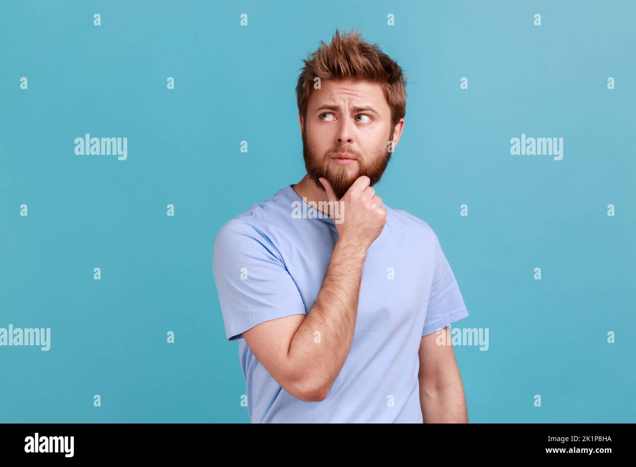 Portrait of unhappy pensive young bearded man keeping hand on chin, looking concentrated into distance, thinking about important things. Indoor studio shot isolated on blue background. Stock Photo