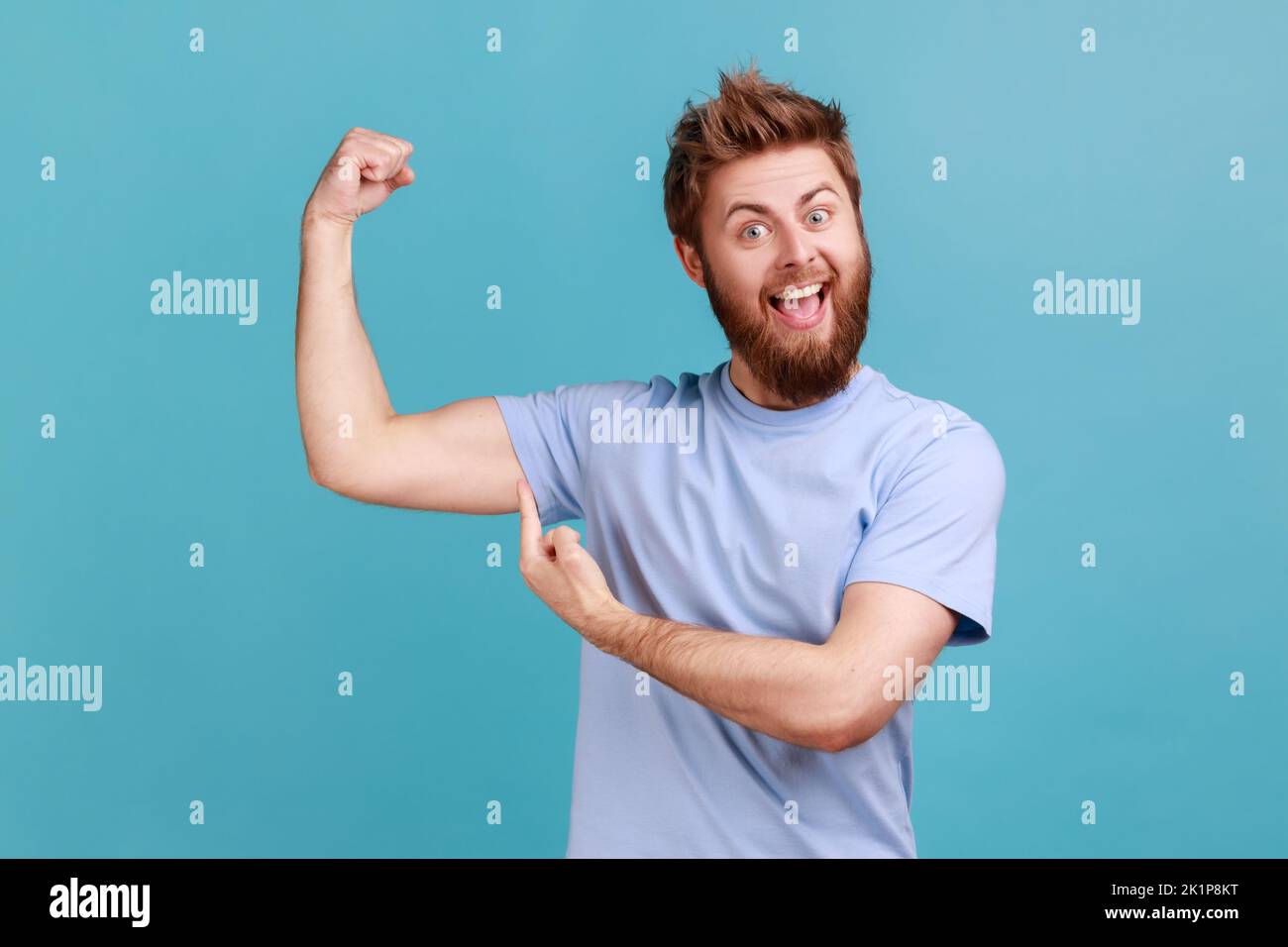Portrait of excited bearded man pointing finger on his arm muscle, pumping in gym, discount, looking at camera with satisfied face. Indoor studio shot isolated on blue background. Stock Photo