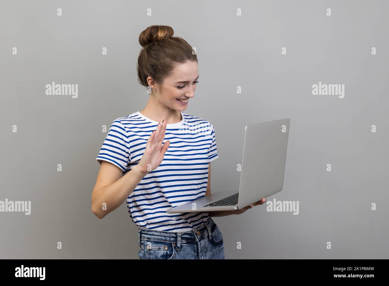 Portrait of friendly woman wearing striped T-shirt standing with laptop, looking at display and waving hand, saying hi, having video call. Indoor studio shot isolated on gray background. Stock Photo