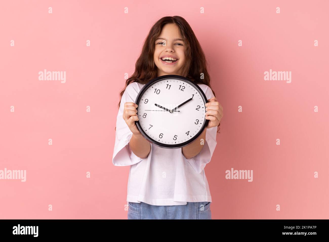 Portrait of smiling delighted little girl wearing white T-shirt holding wall clock, being happy, deadline, satisfied with completed home task. Indoor studio shot isolated on pink background. Stock Photo