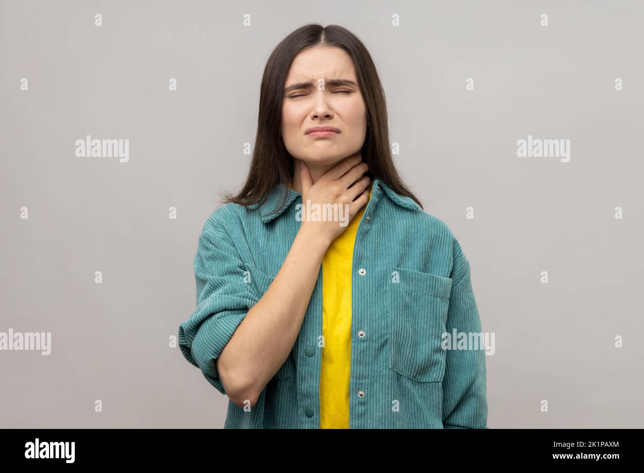 Portrait of sick dark haired woman touching sore neck feeling unwell, suffering tonsils inflammation, medical concept, wearing casual style jacket. Indoor studio shot isolated on gray background. Stock Photo