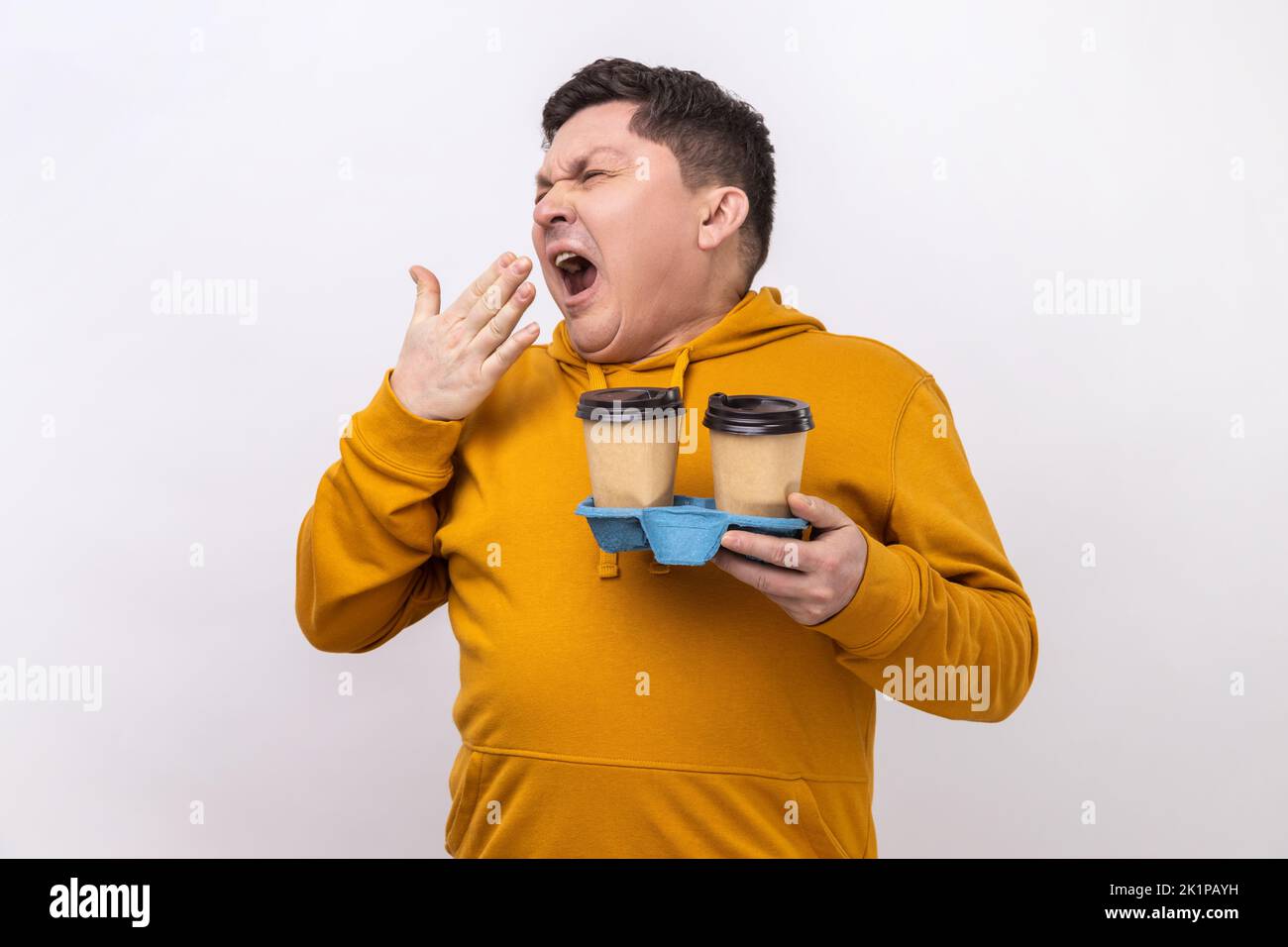 Portrait of tired exhausted man holding coffee cups and yawning, feeling bored after stressful working day, wearing urban style hoodie. Indoor studio shot isolated on white background. Stock Photo