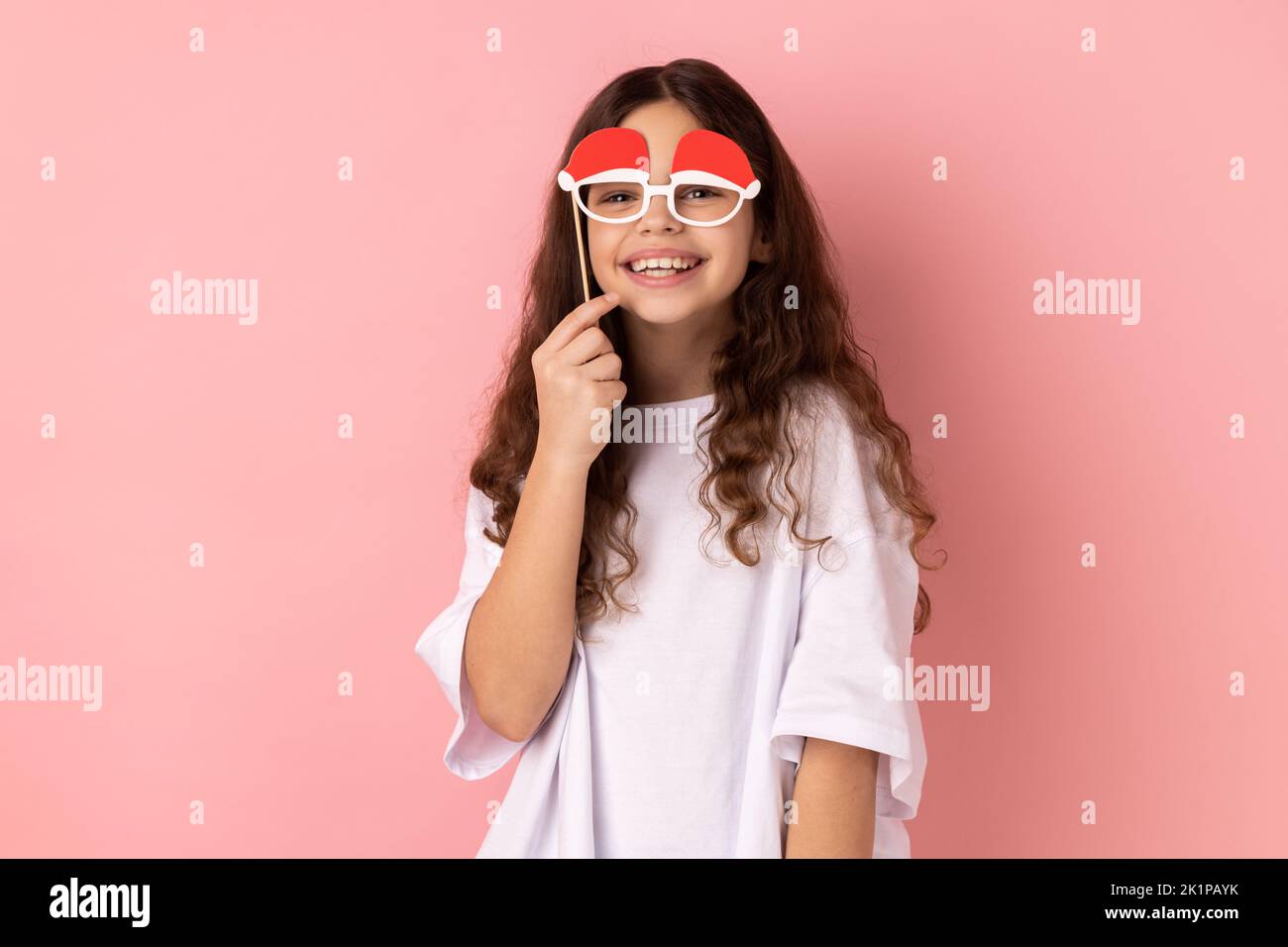 Portrait of little girl wearing white T-shirt holding funny paper glasses, having fun playing game, wearing masquerade accessory. Indoor studio shot isolated on pink background. Stock Photo