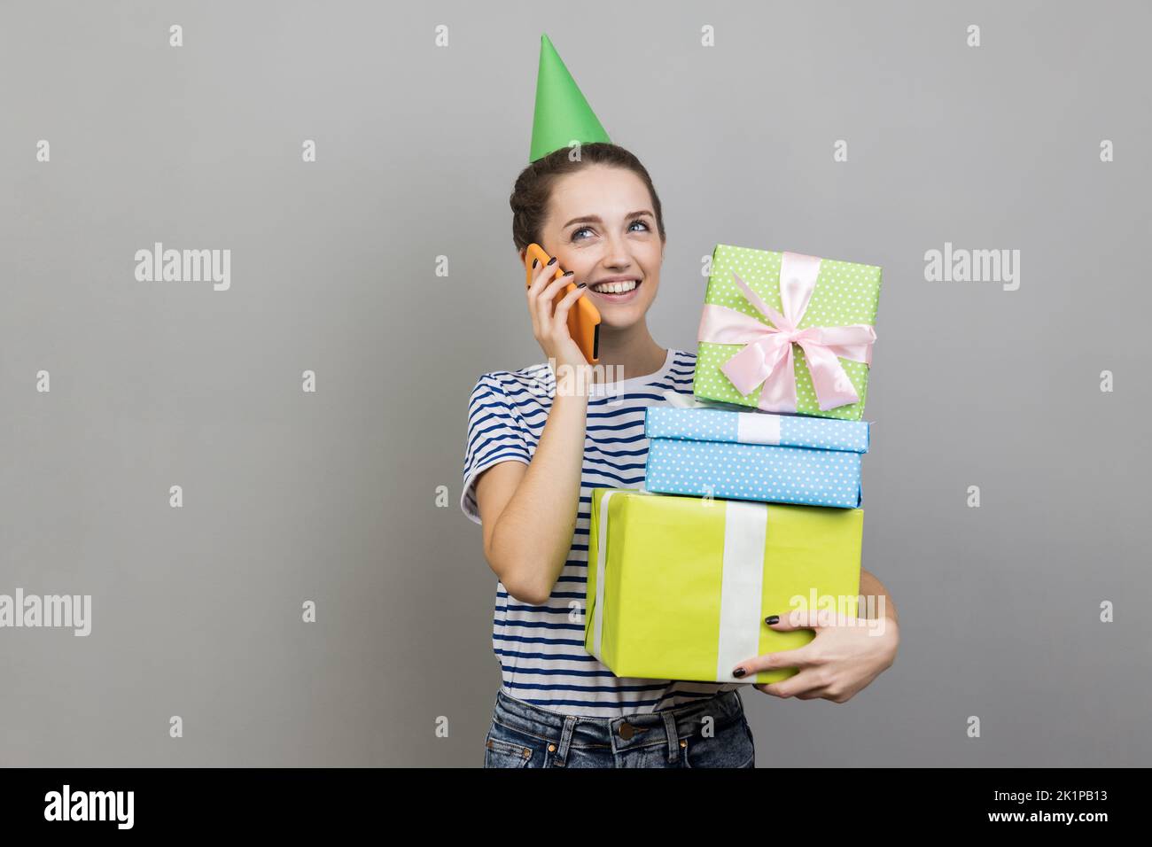 Portrait of optimistic woman wearing striped T-shirt and party cone, holding stack of gift boxes and talking phone, boasting her presents. Indoor studio shot isolated on gray background. Stock Photo